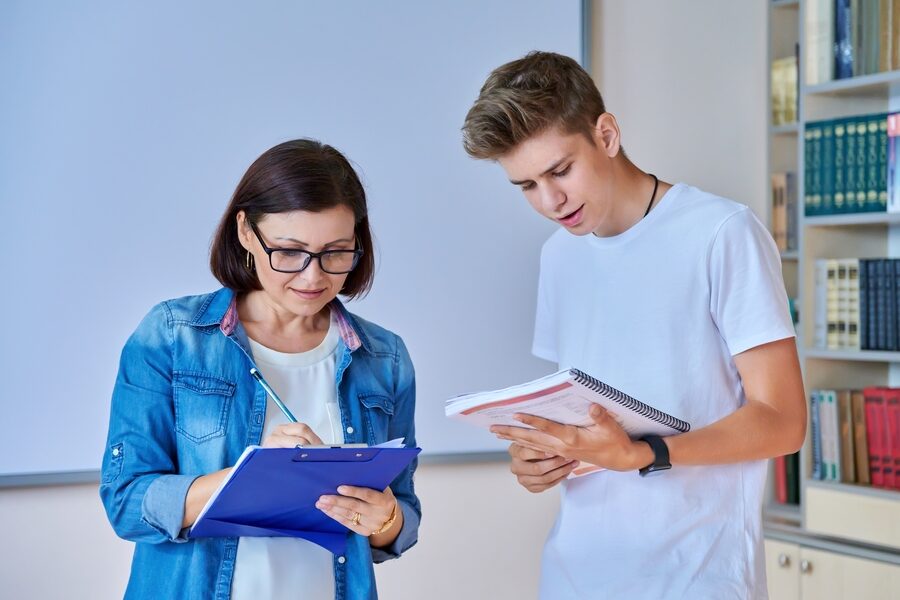 College student guy teenager meeting discussing with teacher mentor in classroom